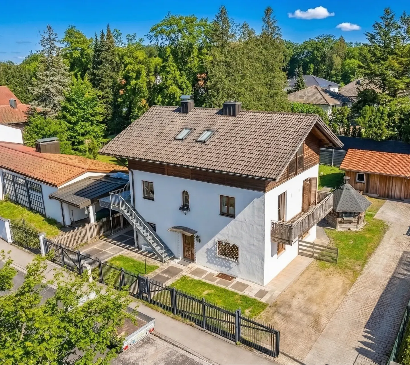 Two-story white house with brown trim and roof, surrounded by green trees and a black fence. A metal staircase leads to the second floor.