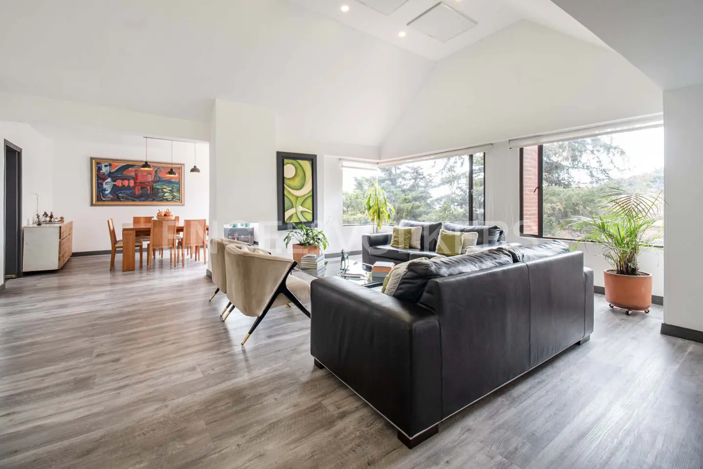 Bright, open-concept living room with gray wood floors, black leather sofas, and large windows overlooking greenery. Dining area visible in background.
