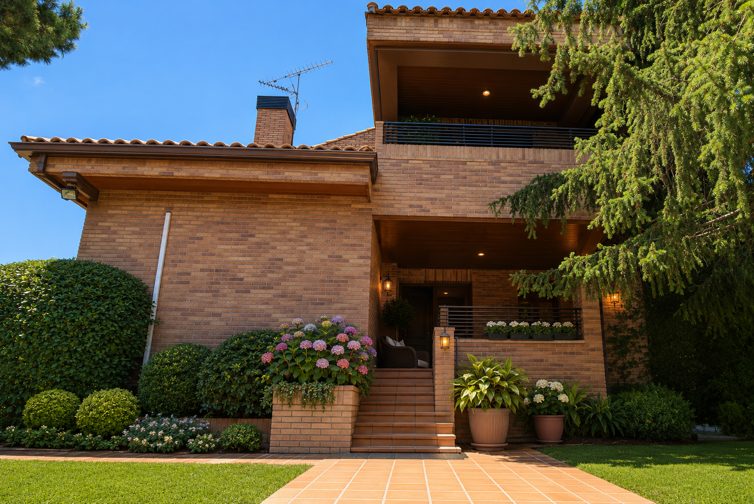Two-story brown brick house with a tiled roof, front steps, balcony, and lush landscaping under a clear blue sky.
