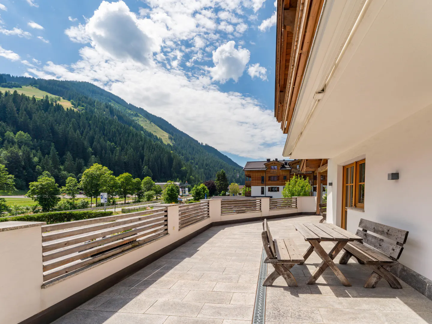 Outdoor patio with wooden table and benches, stone flooring, and a mountain view under a blue sky with white clouds.