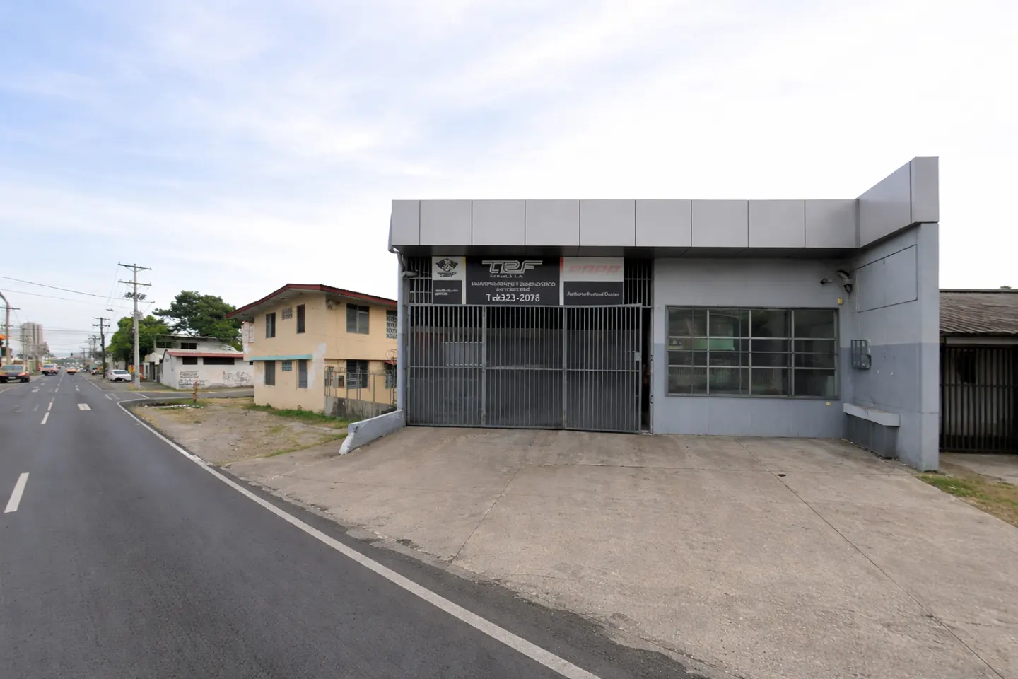 Gray auto shop with a gated entrance and signs, next to a tan two-story house, along a road.
