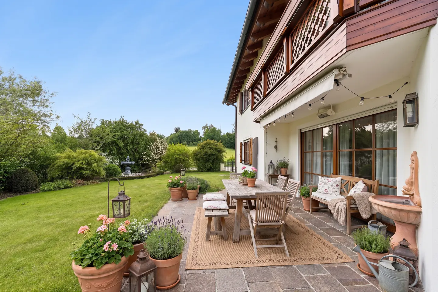 Outdoor patio with a wooden table and chairs on a stone patio, with a green lawn and garden in the background.