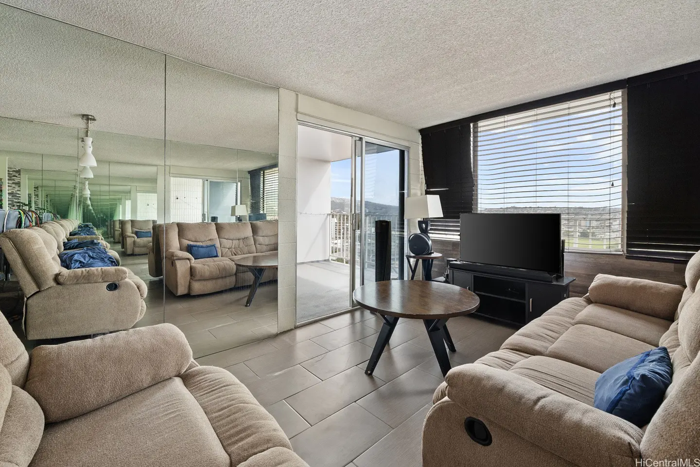 Living room with beige sofas, a round table, and a TV. A mirrored wall creates an infinity effect. Sliding glass doors lead to a balcony.