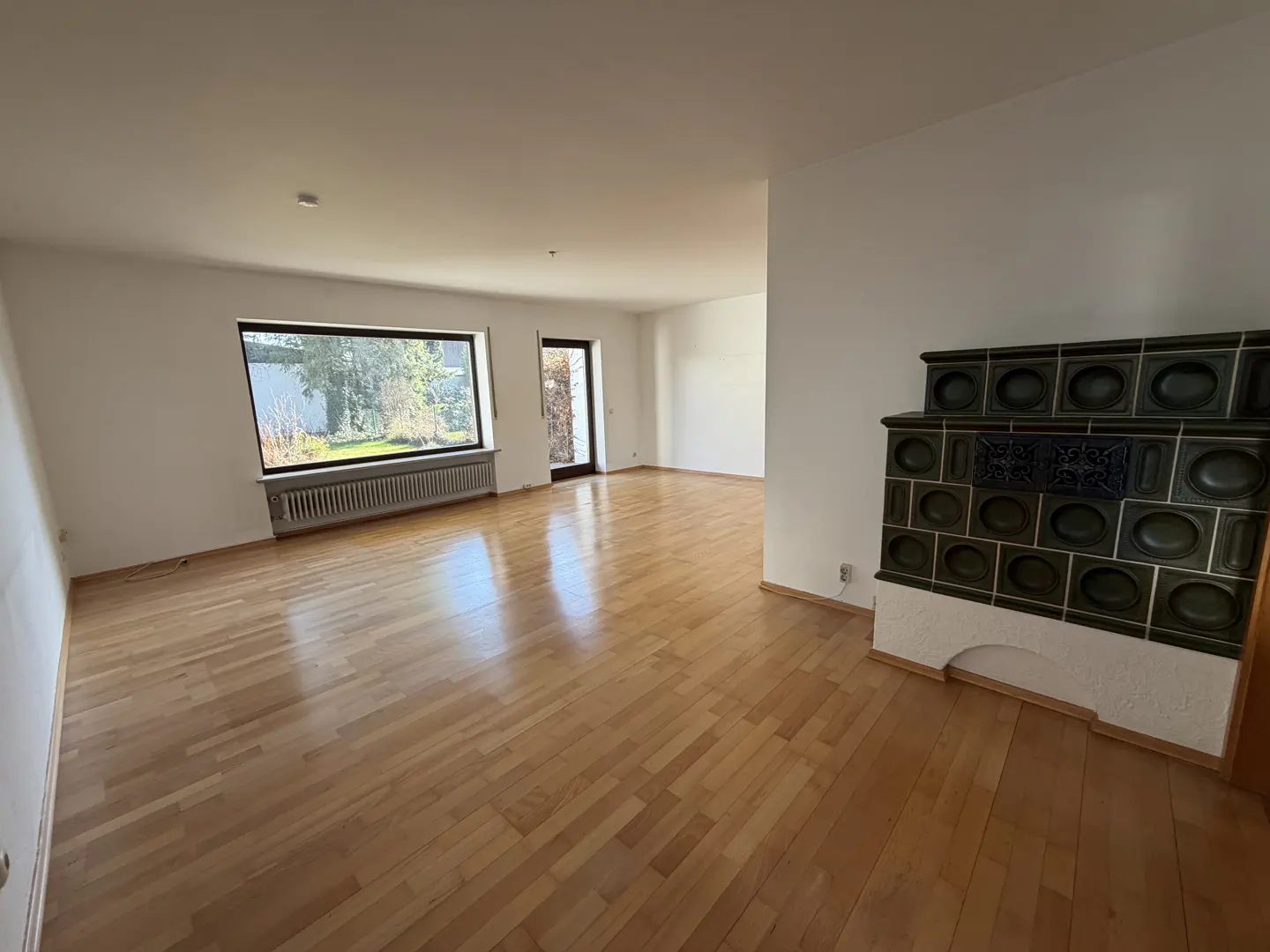 Bright, empty room with wood floors, white walls, large window, radiator, door, and a tiled stove.