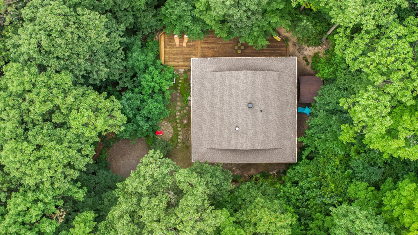 Aerial view of a house with a gray roof surrounded by lush green trees. A wooden deck with furniture is visible.