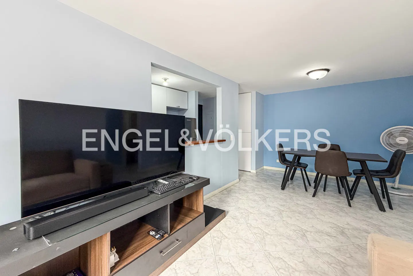 Living room with a large TV, a dining table with four chairs, and a view into the kitchen. The walls are light blue.