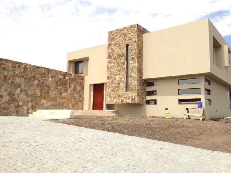 Modern beige house with stone accents, a brown front door, and a stone driveway under a cloudy sky.