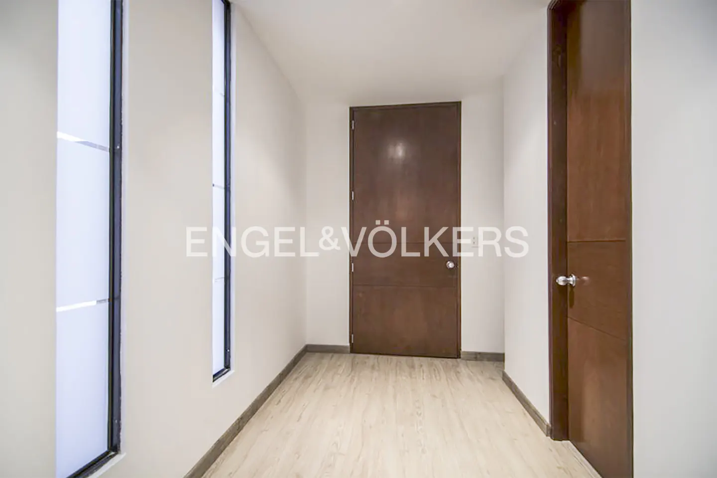 A bright hallway with light wood floors, white walls, two tall narrow windows, and two dark wood doors.