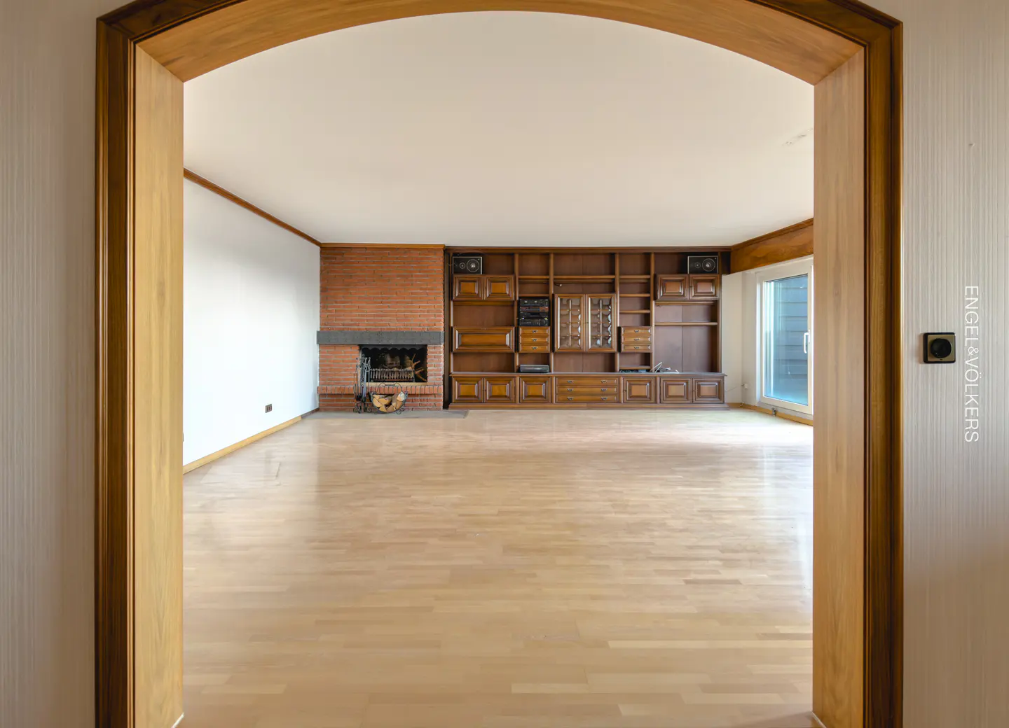 View of a living room through a wooden archway. The room has a brick fireplace, built-in wooden shelves, and light wood floors.