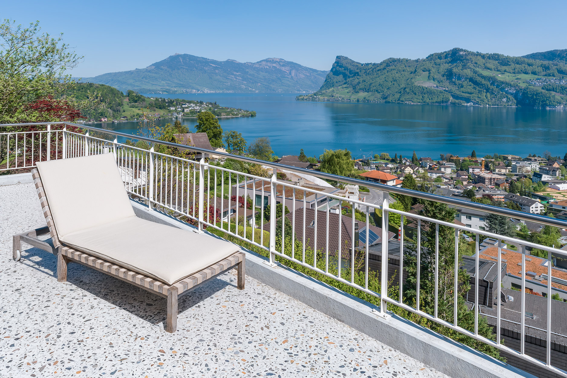 A lounge chair sits on a balcony overlooking a lake, mountains, and a town on a sunny day.