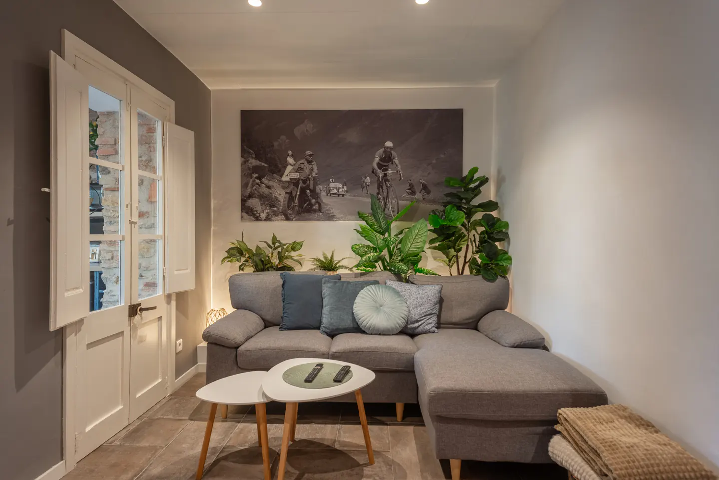 Living room with gray sectional sofa, blue pillows, and two white tables. A black and white photo hangs above the sofa, with plants in front. White doors are on the left.
