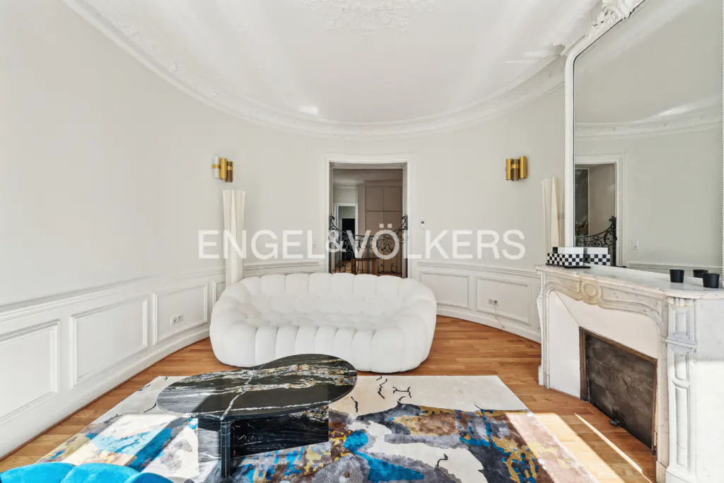 Bright living room with white walls, wood floors, and curved design. A white sofa sits near a black marble table and colorful rug. A fireplace and mirror are on the right.