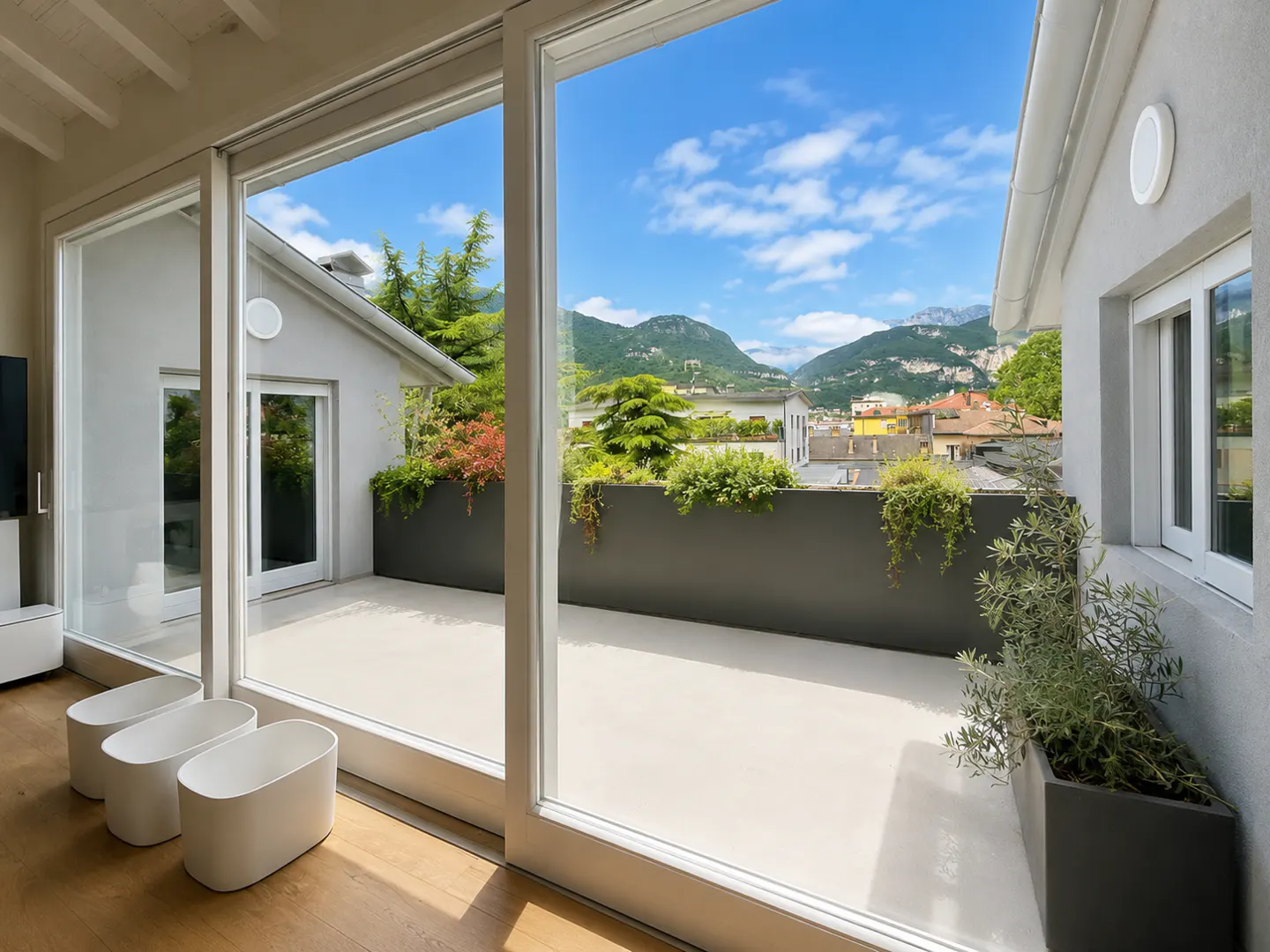 Modern terrace with large glass doors, white planters, and a scenic view of mountains and town under a blue sky.