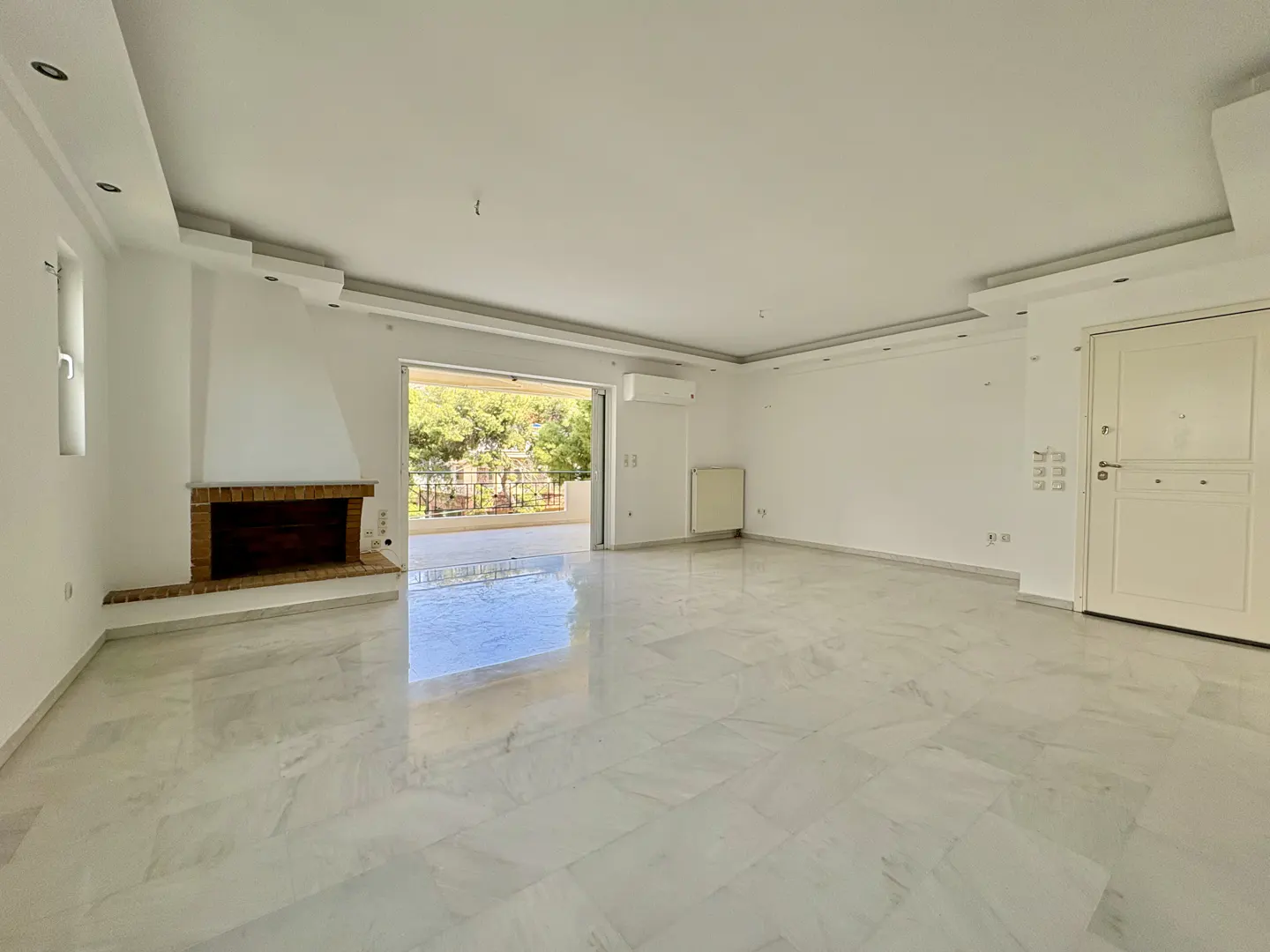 Bright, empty living room with marble floors, white walls, and a brick fireplace. Sliding glass doors lead to a balcony with trees in the background.