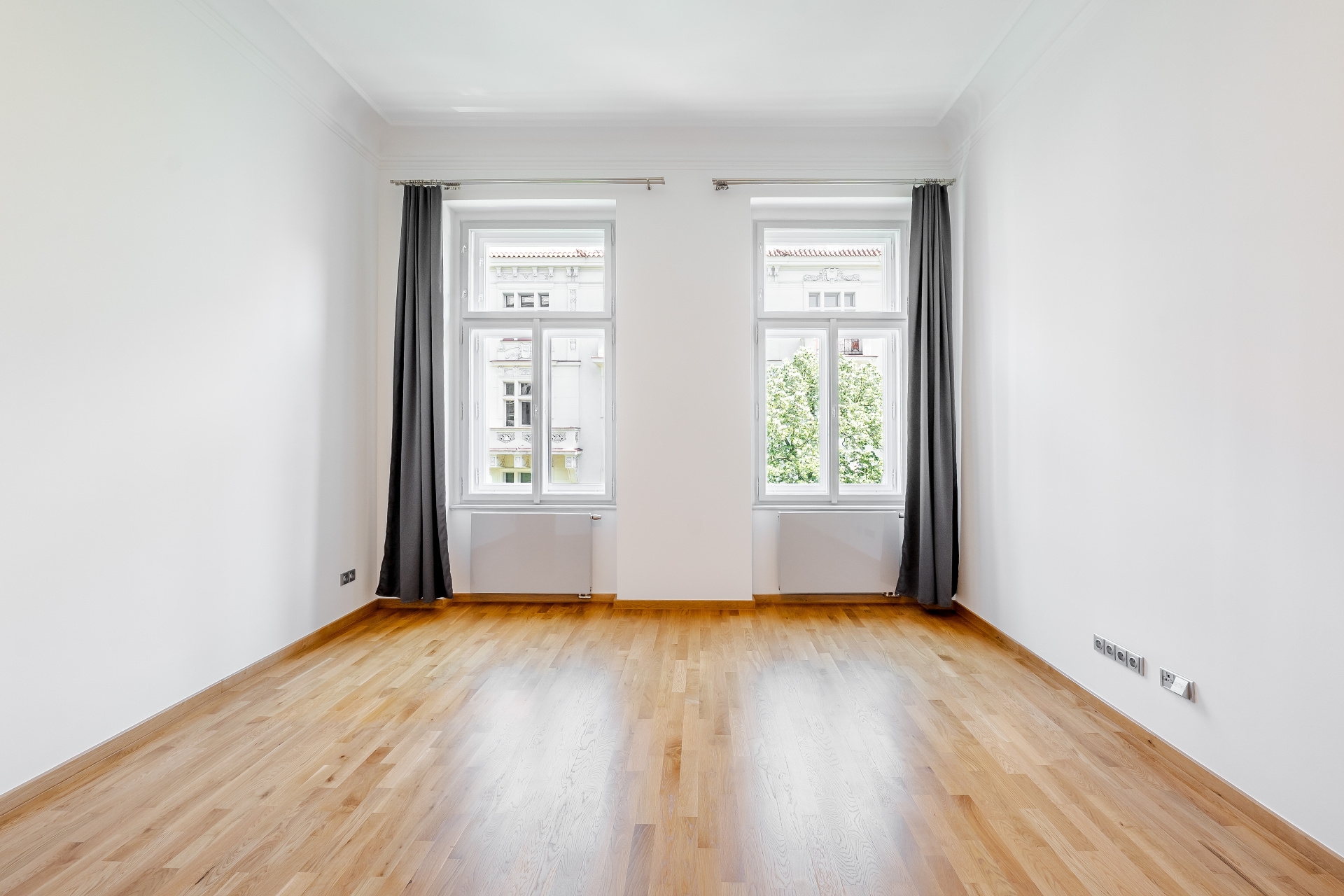 Empty room with hardwood floors, white walls, and two windows with gray curtains. Radiators are under the windows.