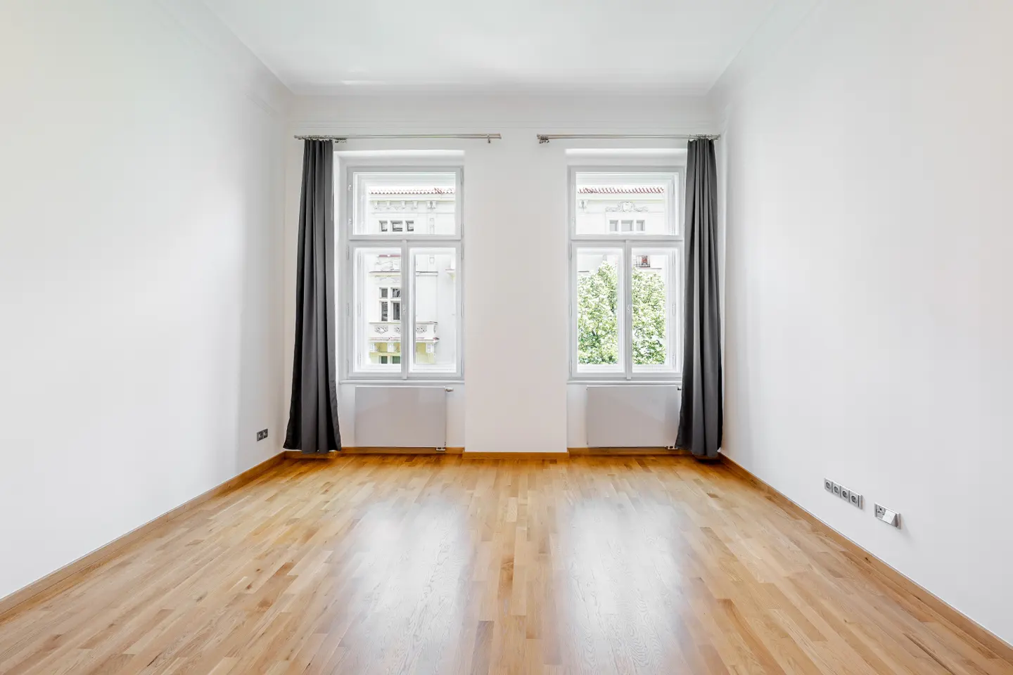 Empty room with hardwood floors, white walls, and two windows with gray curtains. Radiators are under the windows.