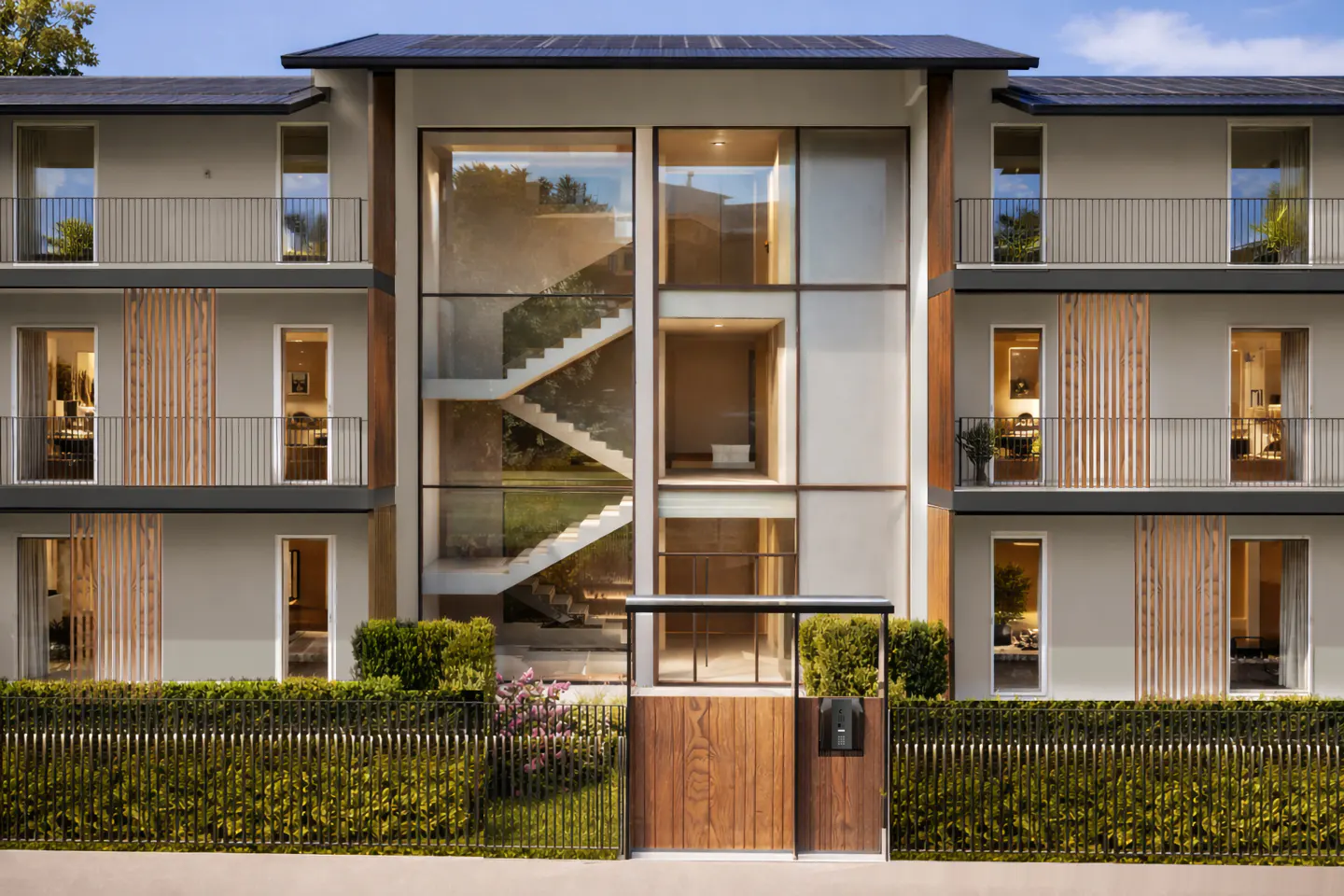 Modern apartment building with a glass-enclosed staircase, balconies, and a wooden gate surrounded by greenery.