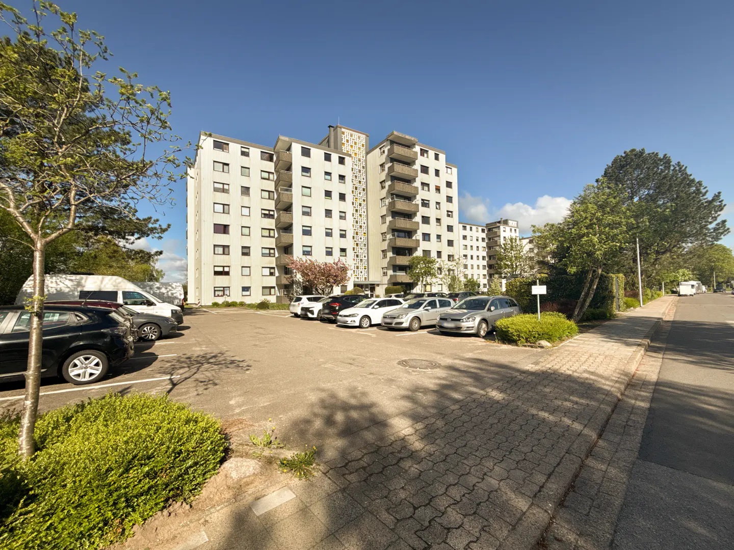 Apartment building with a parking lot full of cars on a sunny day. Trees and bushes surround the building.