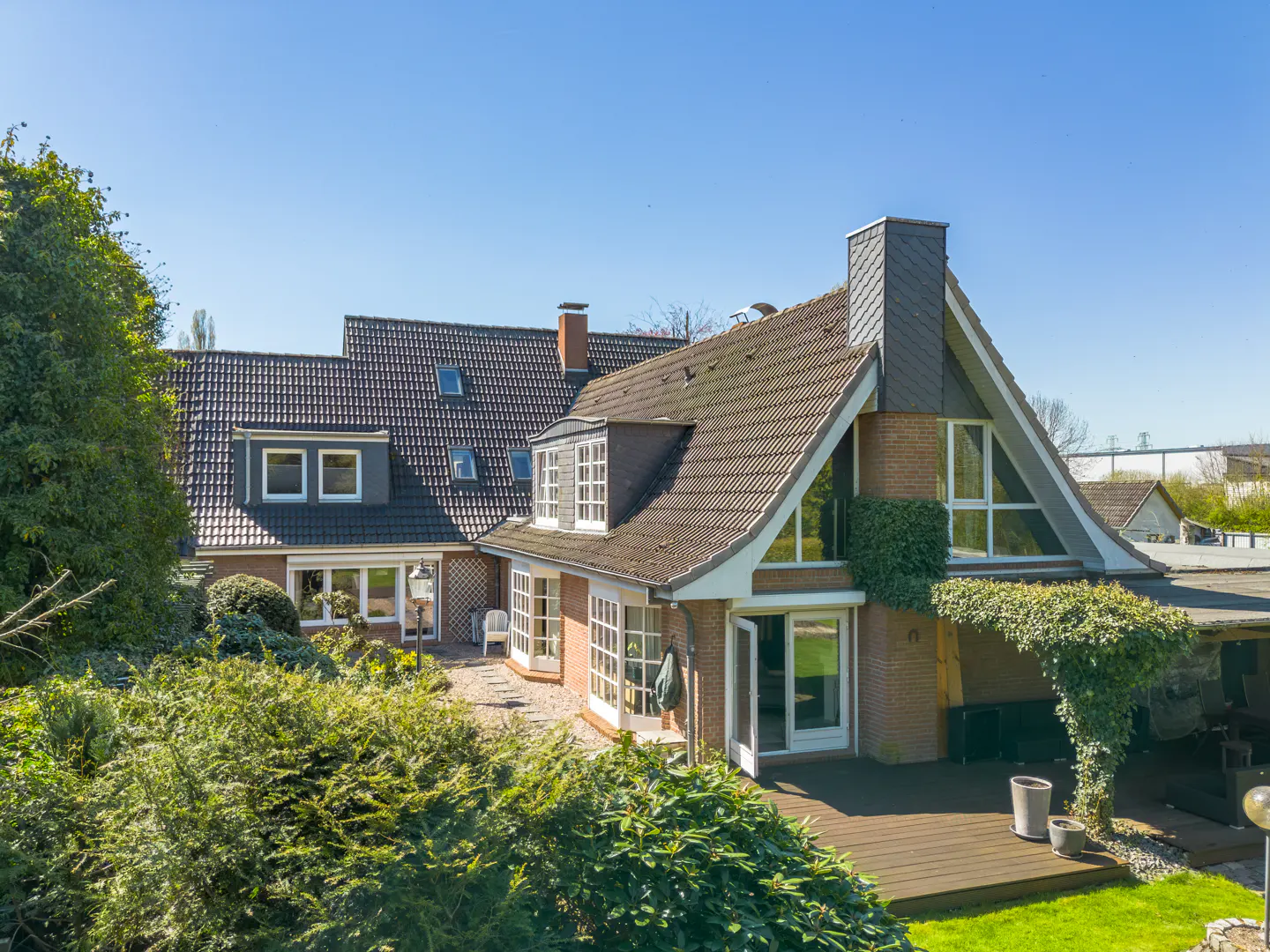 A brick house with a dark tiled roof, large windows, and a wooden deck surrounded by lush green bushes on a sunny day.