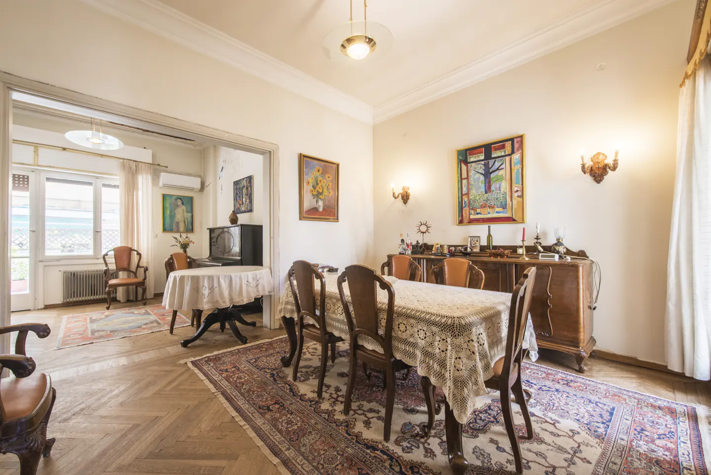 A dining room with a wooden table covered with a white lace tablecloth and wooden chairs. A rug is under the table. There is a wooden cabinet with paintings on the wall.