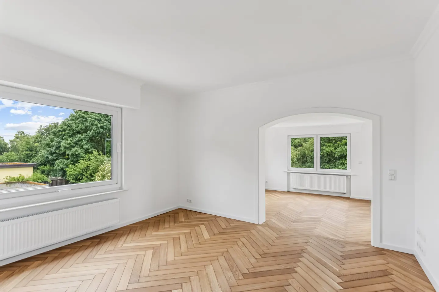Bright, empty room with white walls, herringbone wood floors, and an arched doorway leading to an adjacent room.