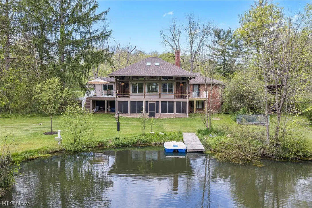 Two-story house with a brown roof, a pond, and a small dock with a boat. The house has a screened-in porch and a deck. Trees surround the house.