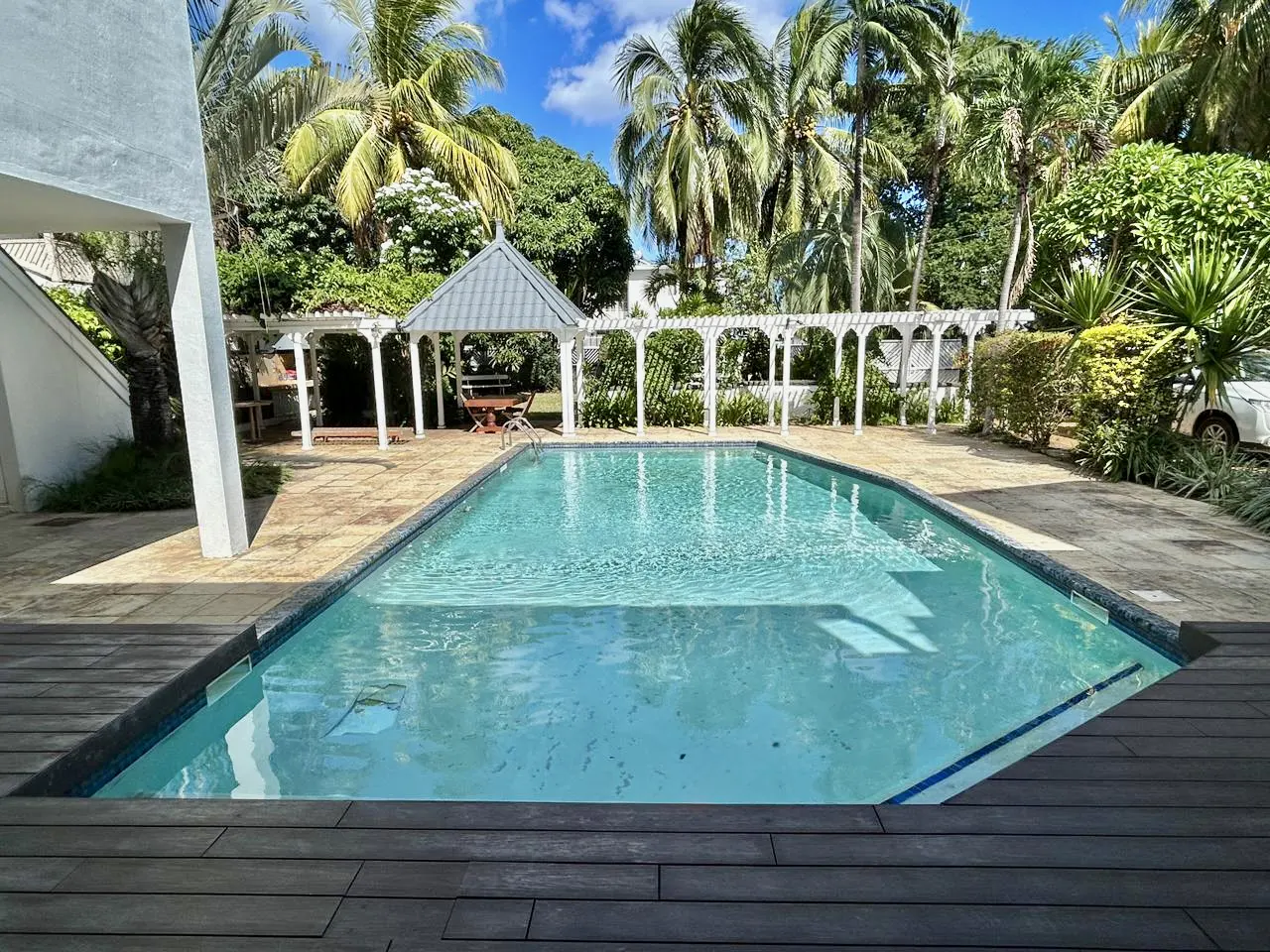 A rectangular pool with clear blue water, surrounded by a wooden deck and a stone patio, with a white gazebo and palm trees in the background.