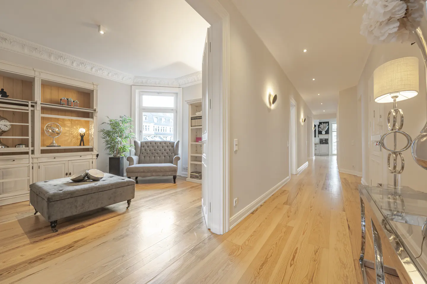 Bright, open-concept living room with hardwood floors, grey ottoman, and armchair. A hallway leads to other rooms.