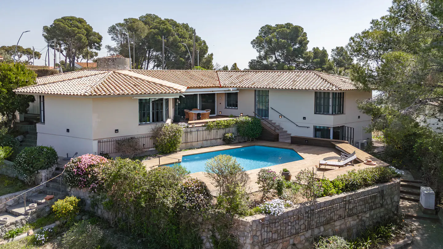 Beige two-story house with a terracotta tile roof, blue pool, patio, and green trees.