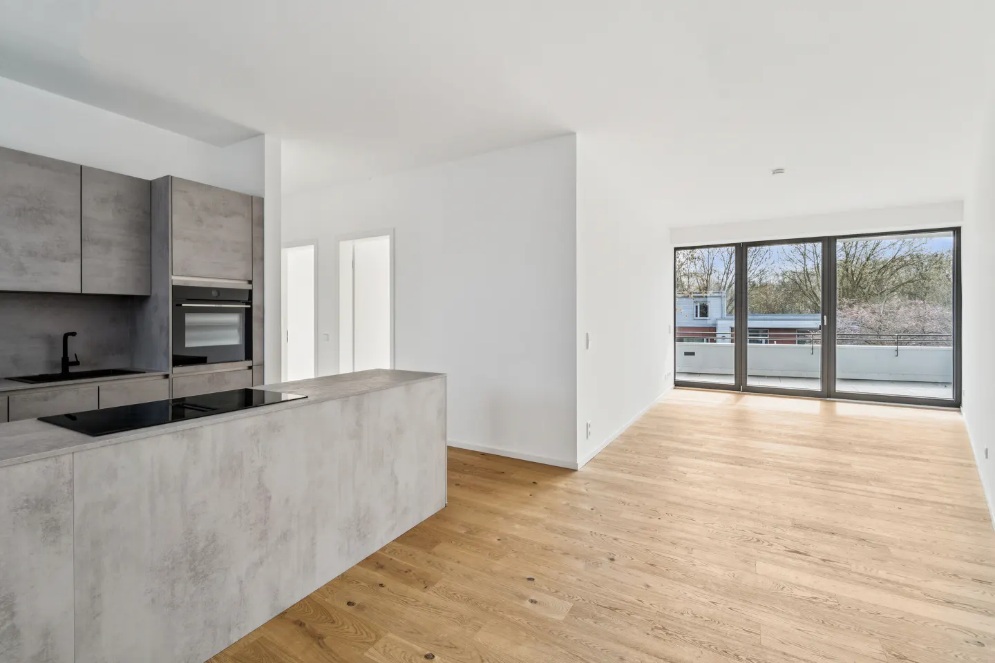 A bright, modern kitchen and living space with gray cabinets, a kitchen island, and wood floors. Sliding glass doors lead to a balcony.