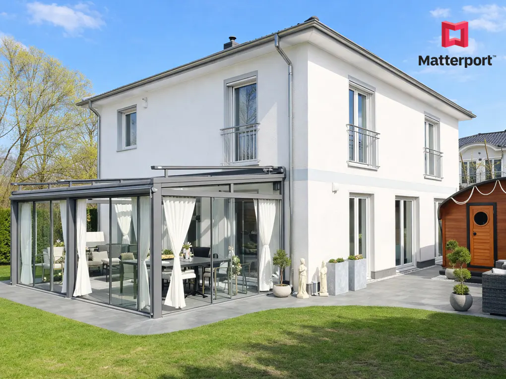 White two-story house with a glass-enclosed patio, white curtains, and a stone patio area on a sunny day.
