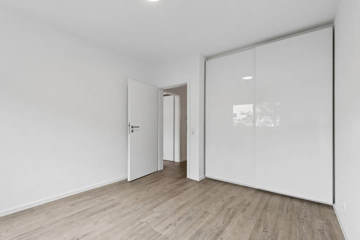 Minimalist room with light wood floors, white walls, a large white sliding-door wardrobe, and an open doorway.