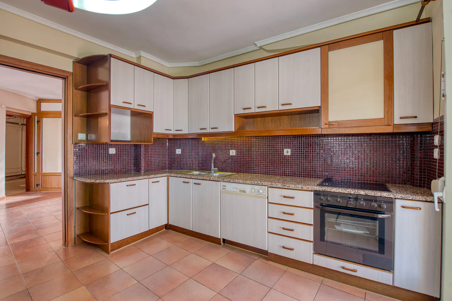A kitchen with white cabinets, brown trim, and a red tile backsplash. A doorway leads to another room with a tiled floor.