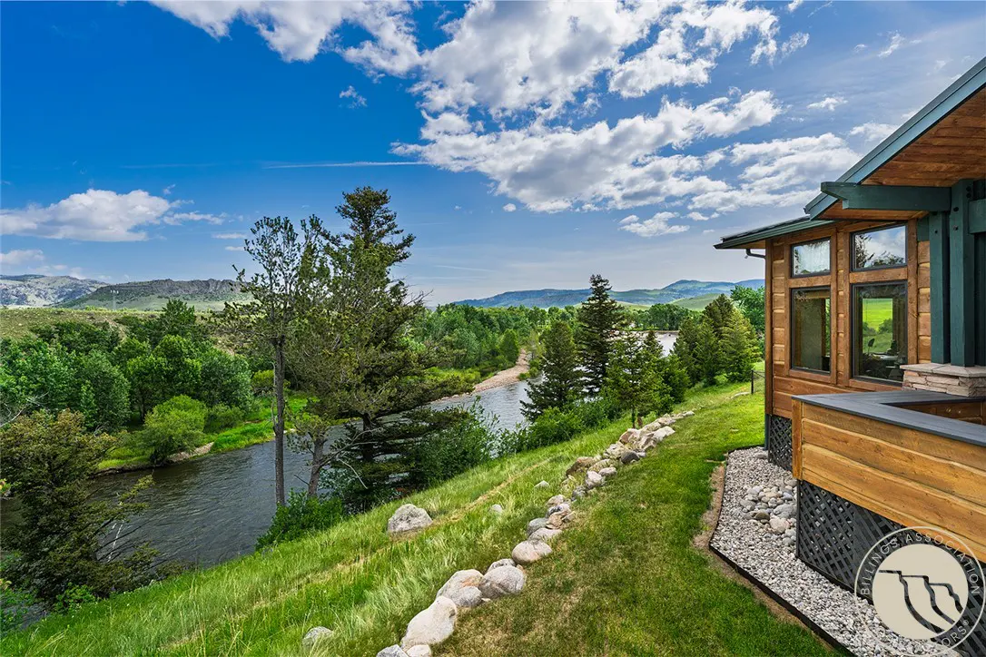Exterior view of a wood-sided house with a river and trees in the background under a blue sky with clouds.