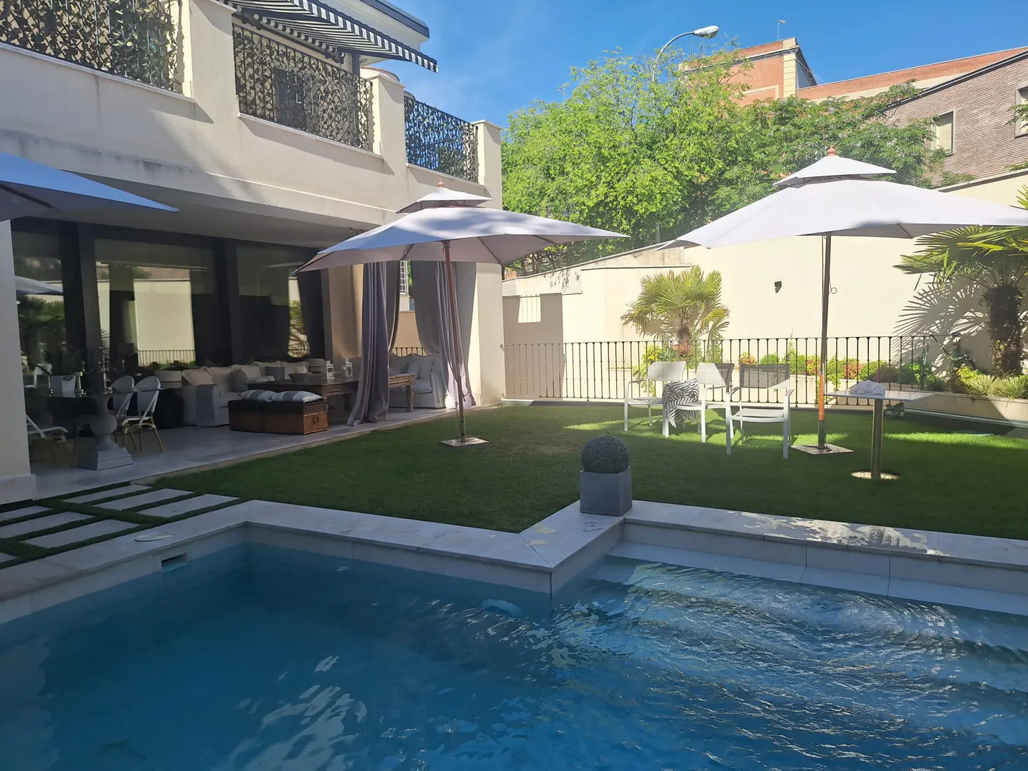 A backyard with a pool, green grass, and two white umbrellas. A patio with white furniture is visible.