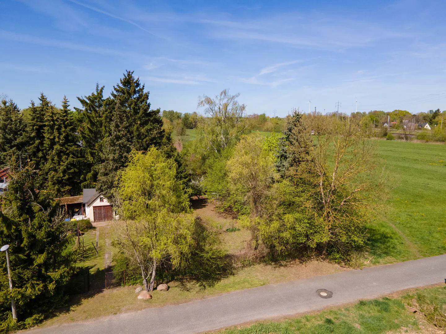 Aerial view of a property with a small house surrounded by trees, a road, and a green field under a blue sky.