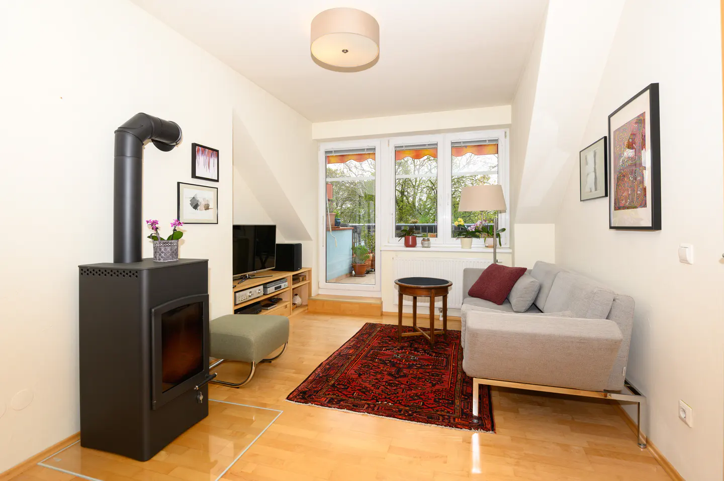 Cozy living room with a black wood-burning stove, gray sofa, and red rug on a light wood floor. Balcony doors let in natural light.