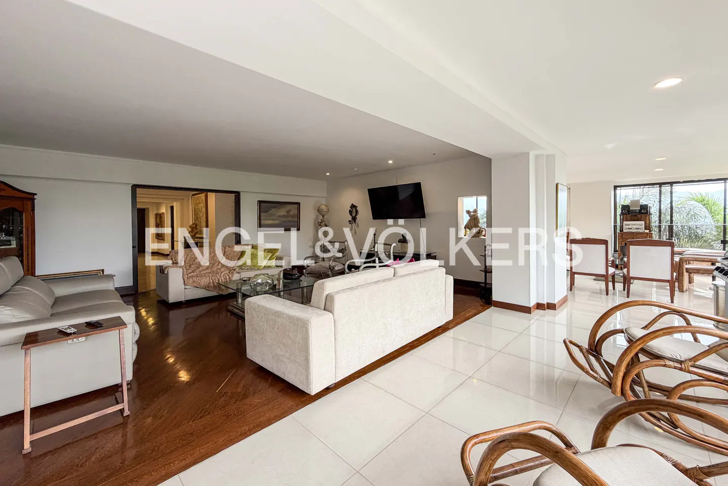 Open-concept living room with white sofas, wood floors, and tile. Large windows offer natural light. Engel & Volkers logo overlay.