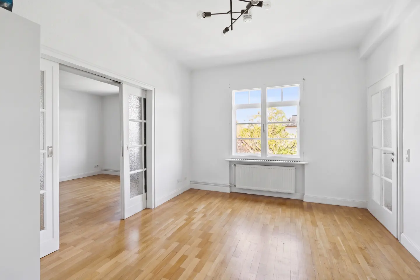 Bright, empty room with wood floors, white walls, and a window showing trees. A modern light fixture hangs from the ceiling.