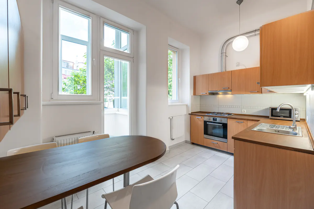 Bright kitchen with light wood cabinets, dark oval dining table, white chairs, stainless appliances, and large windows.