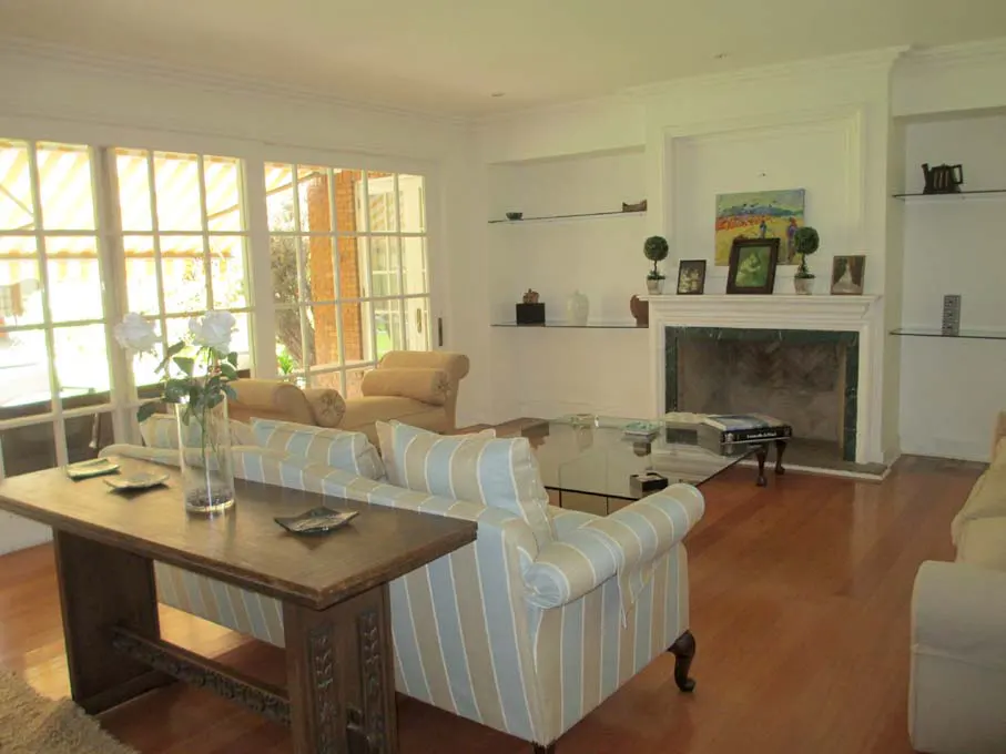 Living room with hardwood floors, fireplace, and large windows. A striped sofa and wooden table are in the foreground.