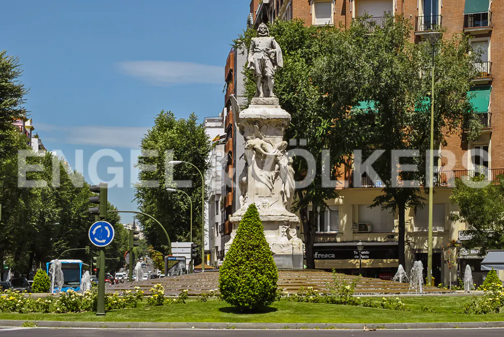 Cityscape view of a roundabout with a statue in the center, surrounded by trees, buildings, and a blue sky.