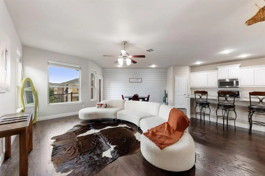 Open-concept living space with a white curved sofa, cowhide rug, and kitchen with bar seating. Dark wood floors and white cabinets.