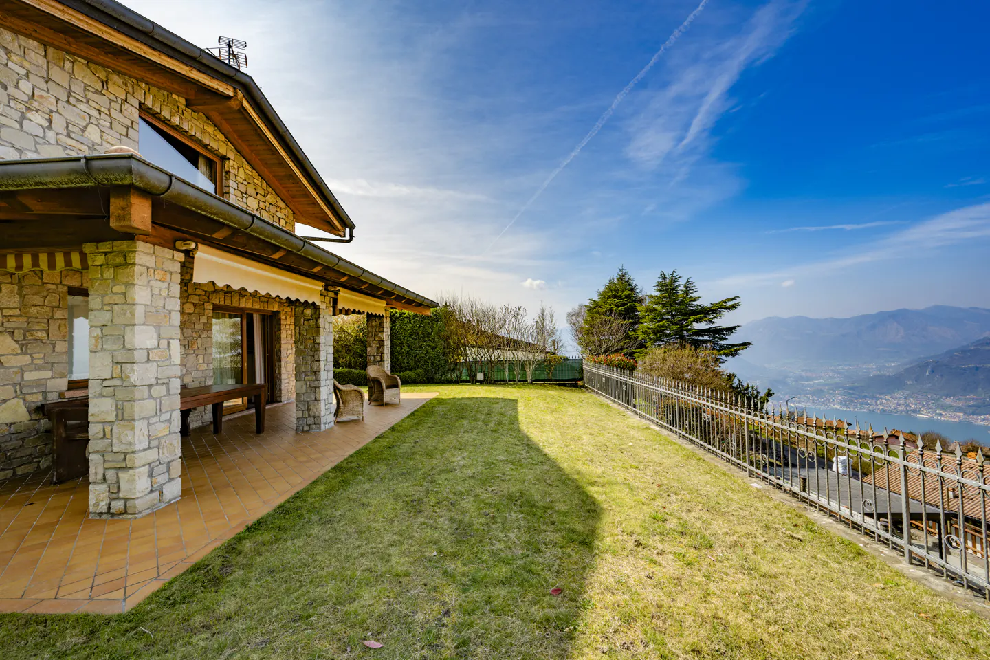 Stone house with a tiled patio, green lawn, and a view of mountains and a lake under a blue sky.