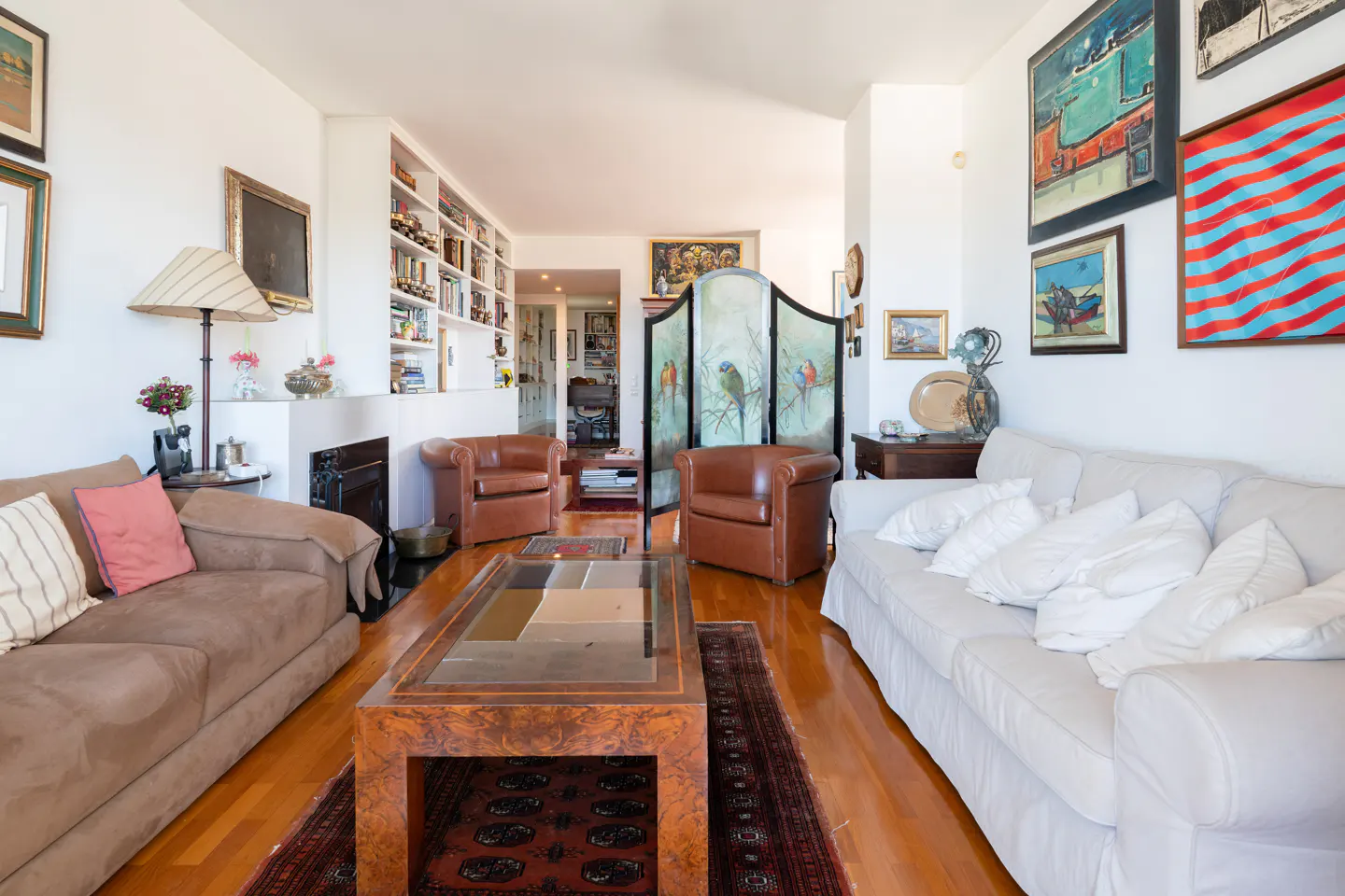 Bright living room with hardwood floors, brown and white sofas, leather chairs, and a large coffee table. Artwork adorns the walls.