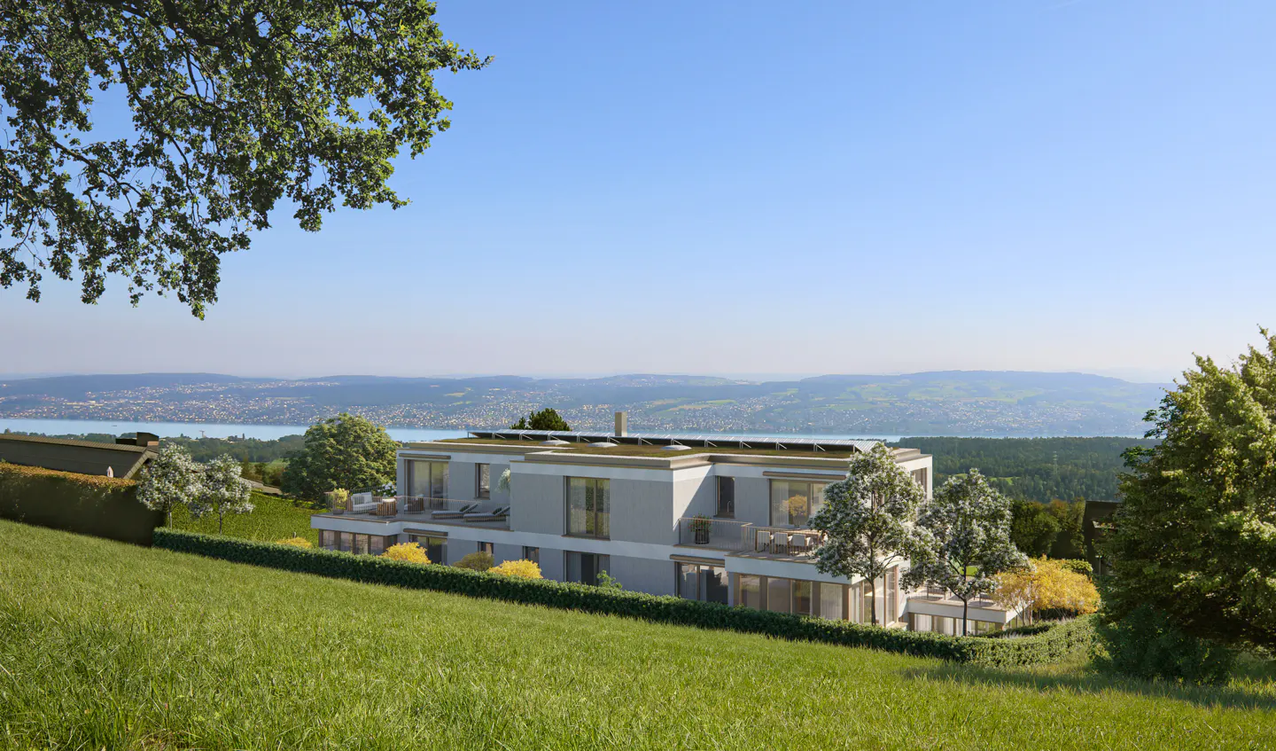 Modern two-story house with balconies, green roof, and solar panels on a grassy hill overlooking a lake and mountains.