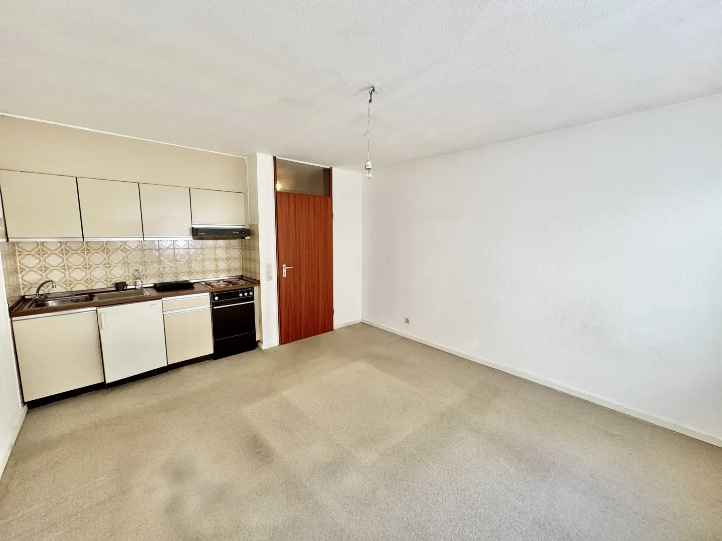 A wide shot of a studio apartment with a kitchen, brown door, and beige carpet.