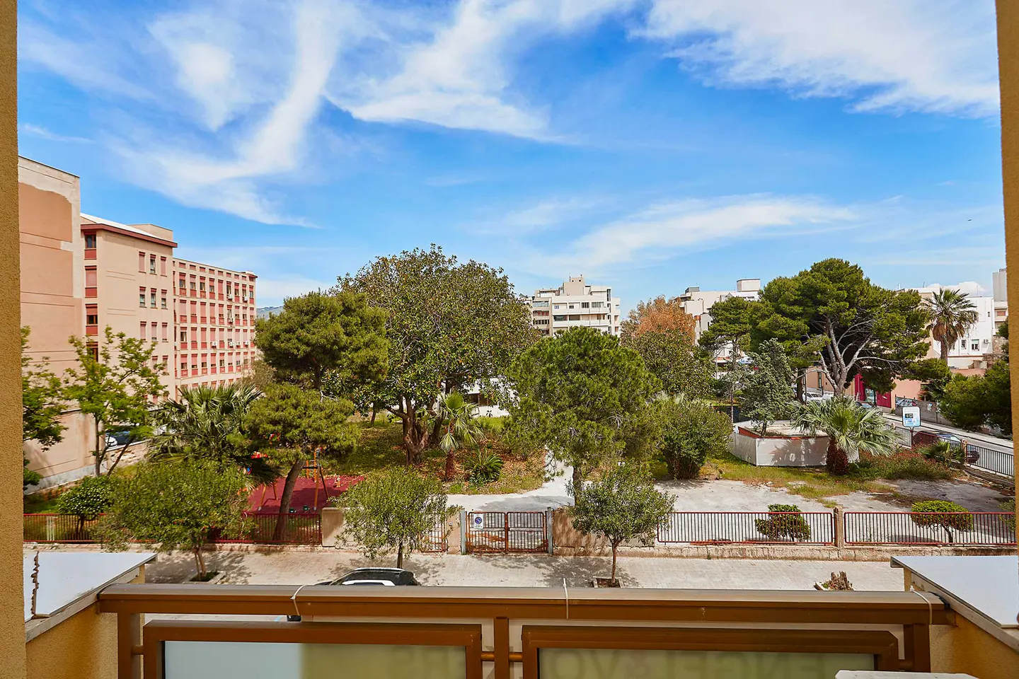 View from a balcony overlooking a park with trees, buildings, and a blue sky with white clouds.