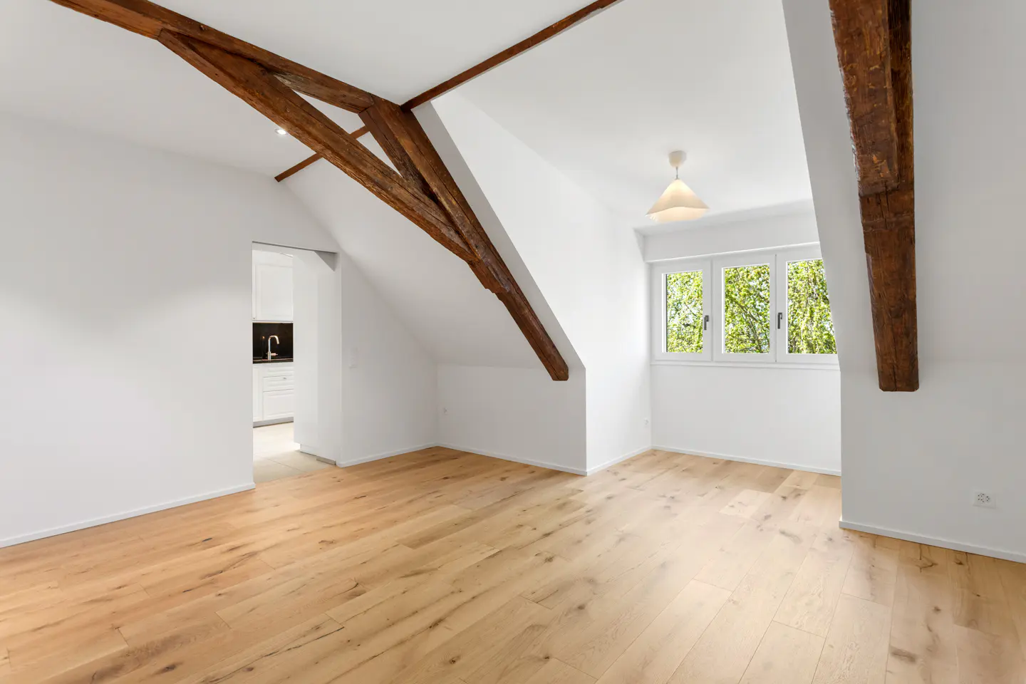 Bright attic room with wood floors, white walls, and exposed beams. A window overlooks green trees. A doorway leads to a kitchen.