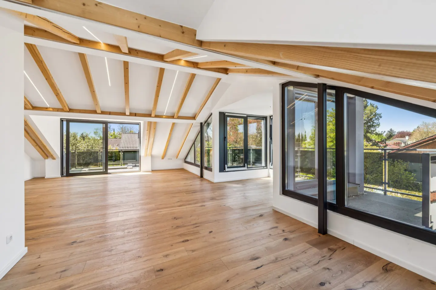 Bright attic room with wood floors, white walls, and exposed wood beams. Black-framed windows offer outdoor views.